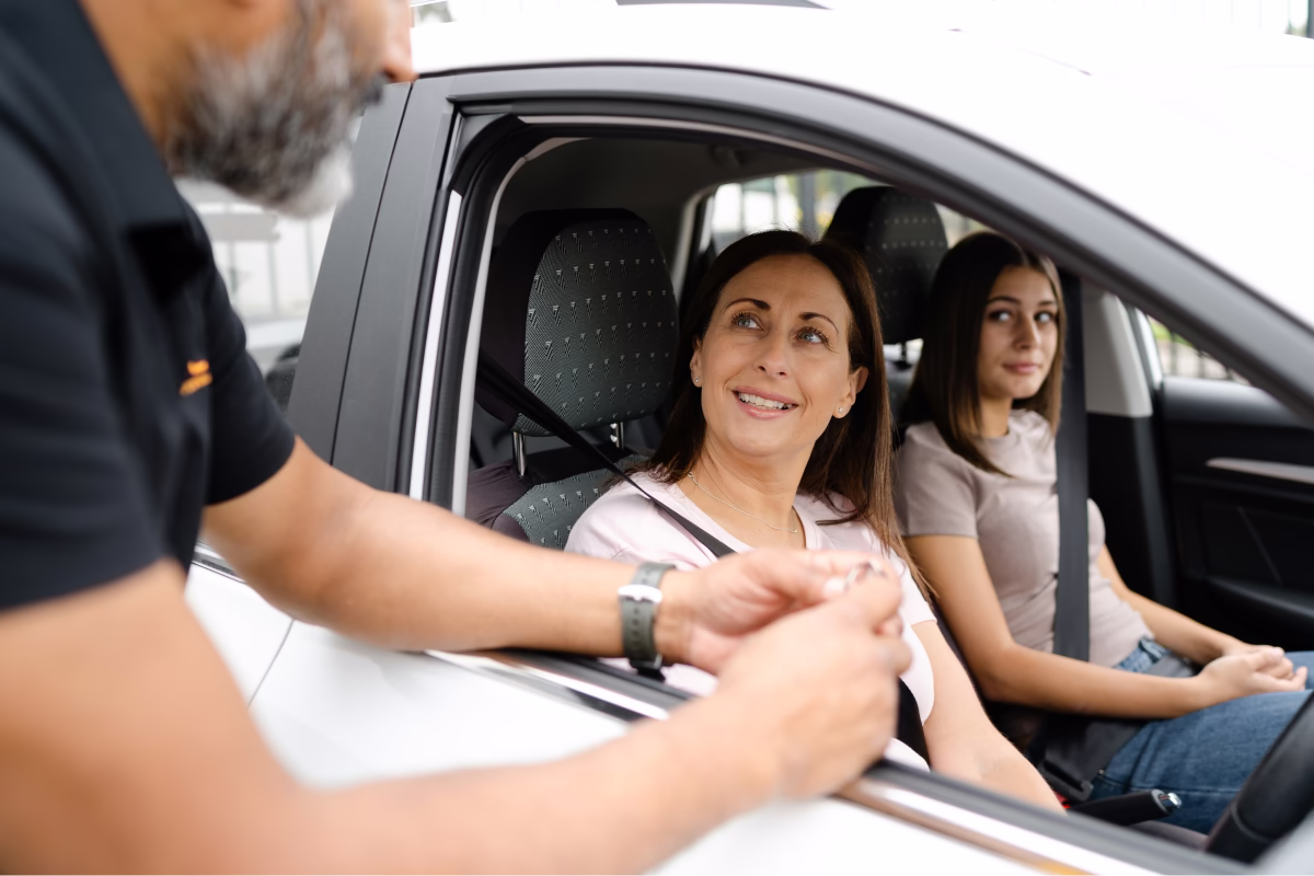 A woman sitting in the driver’s seat of a white car speaks with a man standing outside the window, while a younger woman sits in the passenger seat.