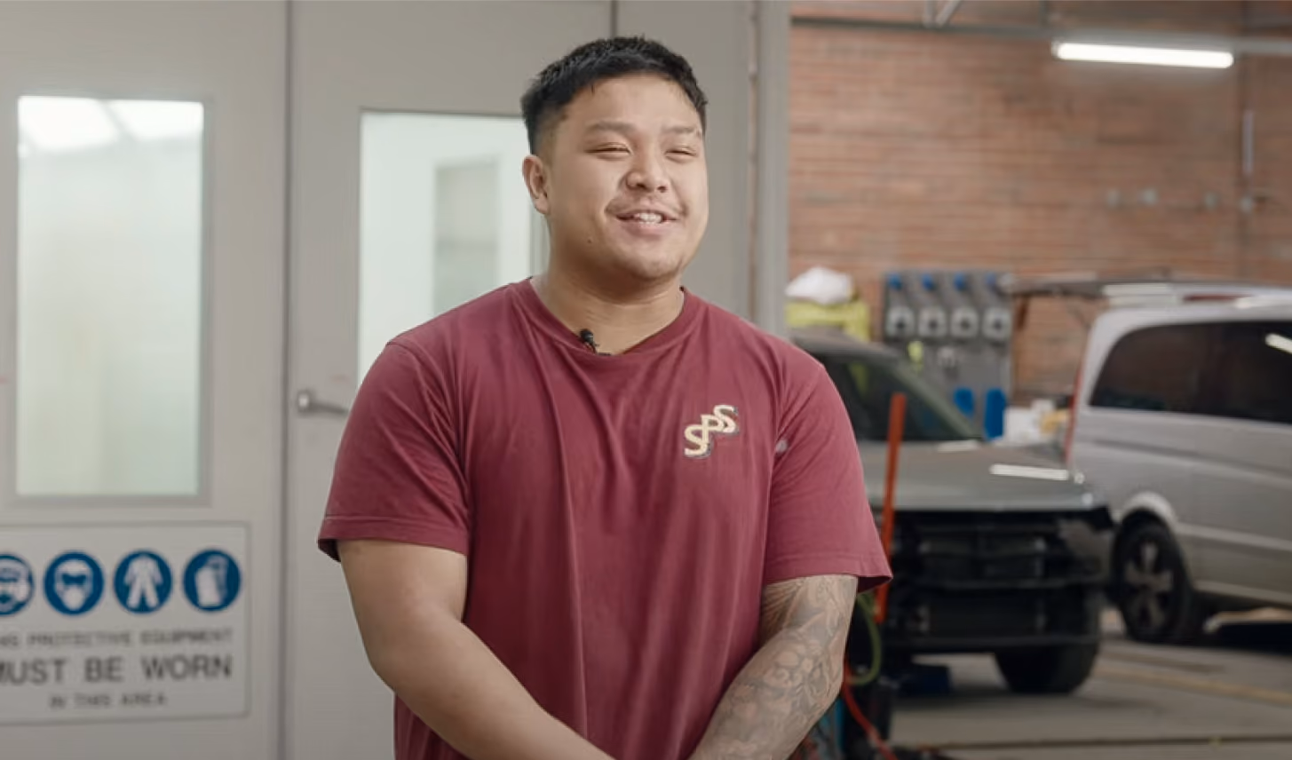 Young man with short hair wearing a maroon t-shirt standing in a workshop or garage, smiling with eyes closed, with vehicles and equipment in the background.
