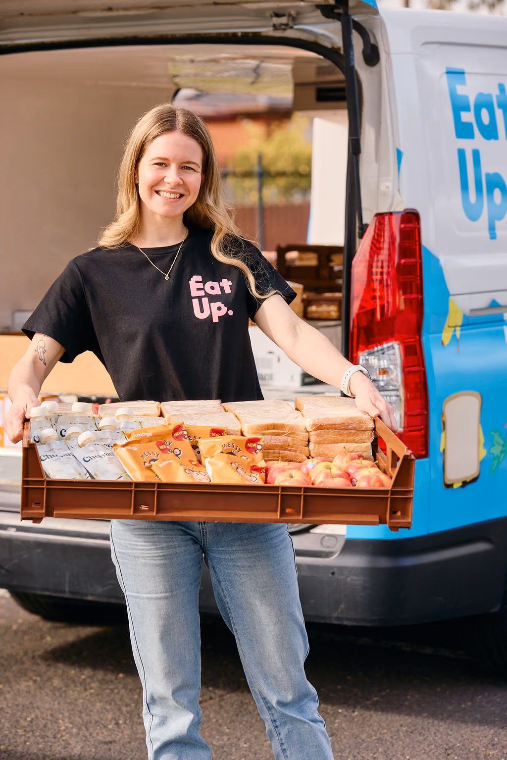 Smiling young woman wearing a black Eat Up t-shirt holding a large tray filled with sandwiches, packaged snacks, and apples, standing in front of a van with Eat Up branding.