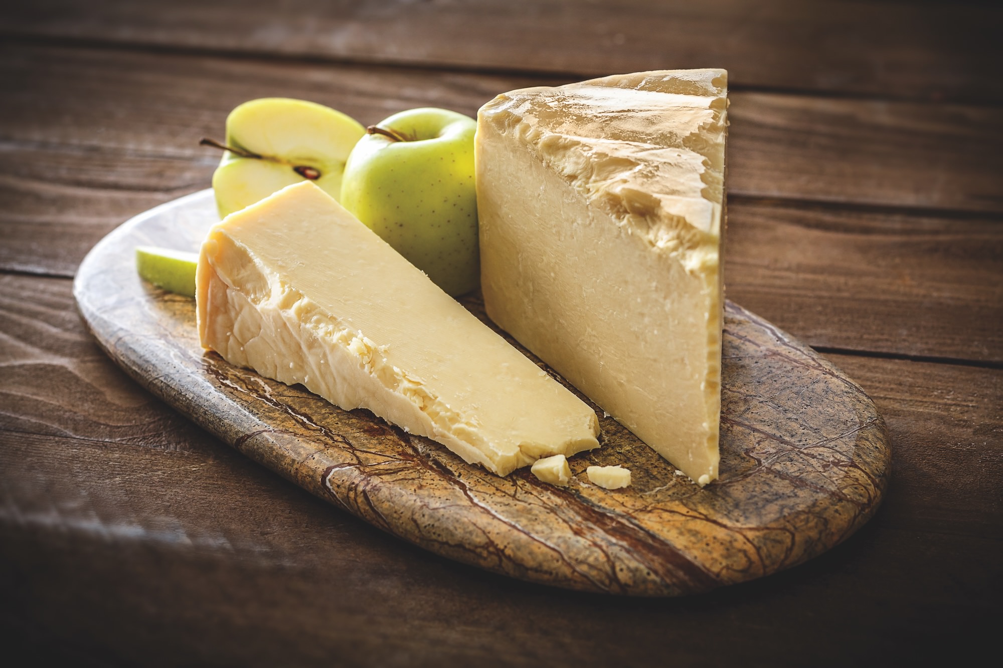 A beautiful wedge of white, raw-milk cheddar atop a rustic cheeseboard with bright green apples in the background.