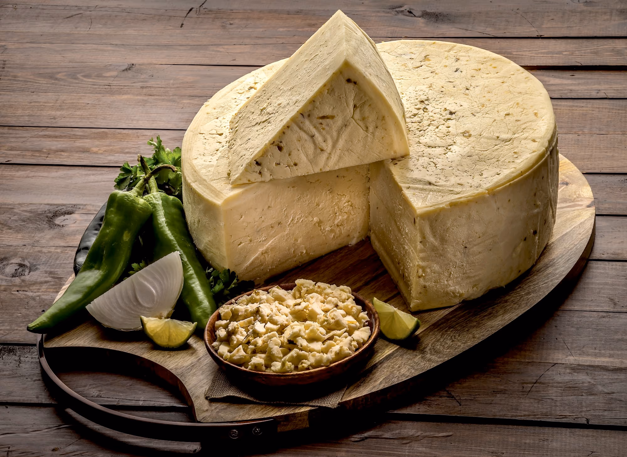 A wheel of queso fresco verde atop a wooden cutting board surrounded by limes, onions, cilantro, and green peppers.  