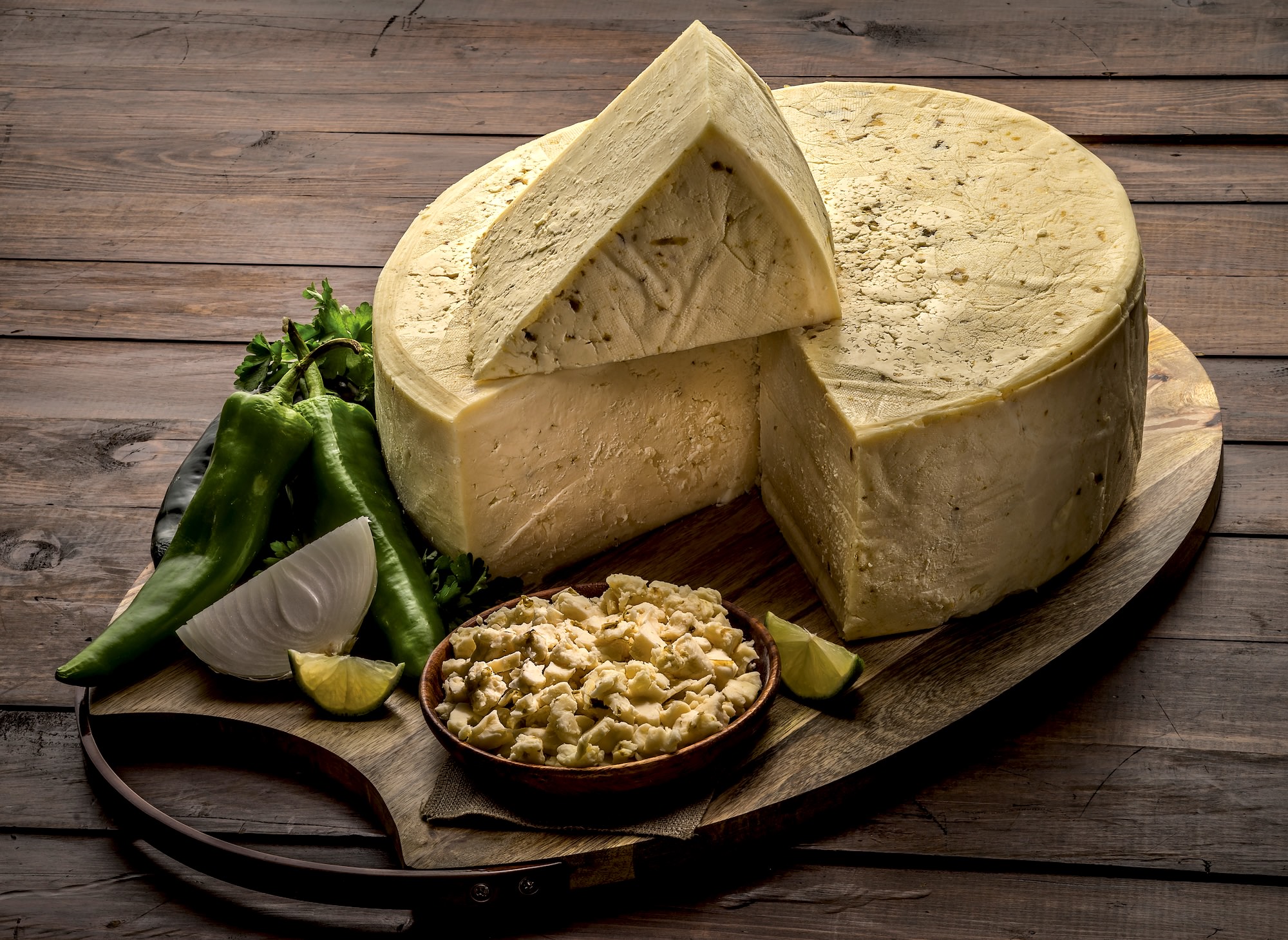 A wheel of queso fresco verde atop a wooden cutting board surrounded by limes, onions, cilantro, and green peppers.  