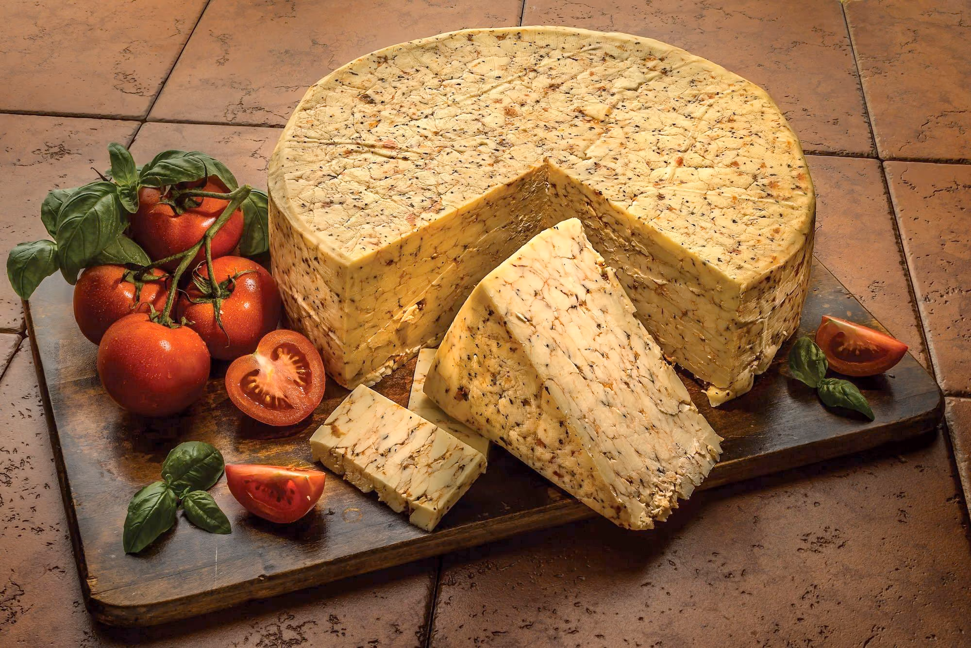 A wheel of tomato-basil flavored cheese on a wooden cutting board surrounded by fresh tomatoes and basil.