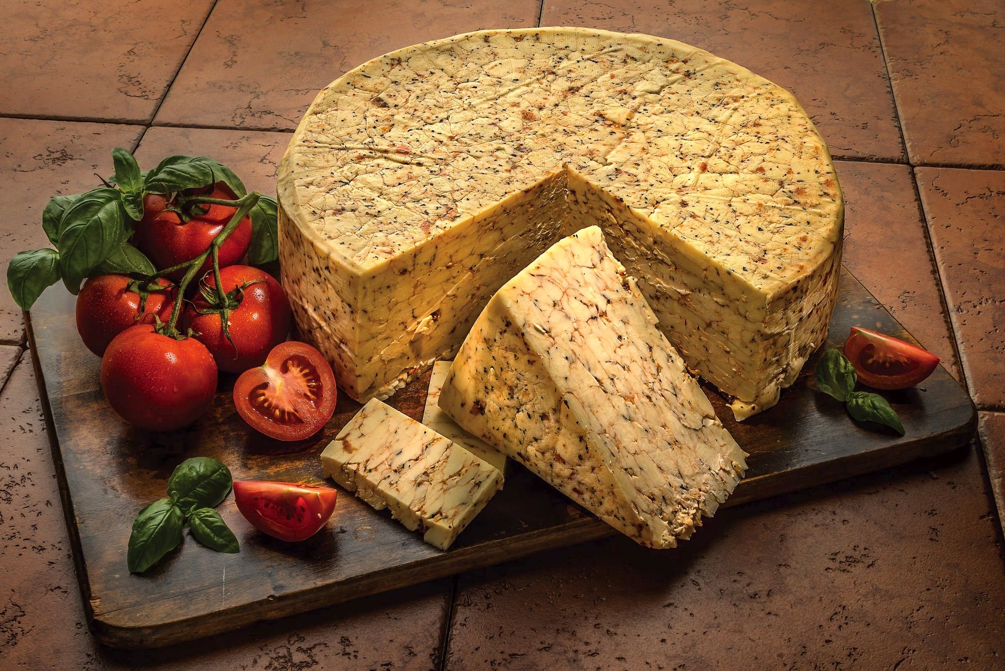 A wheel of tomato-basil flavored cheese on a wooden cutting board surrounded by fresh tomatoes and basil. 