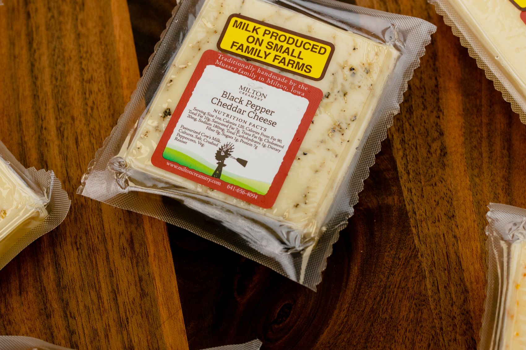 Pale ivory in color and speckled with coarse black pepper throughout, clearly visible through the packaging. The composition highlights the handcrafted, small-batch nature of the cheeses and emphasizes variety, texture, and artisanal quality.