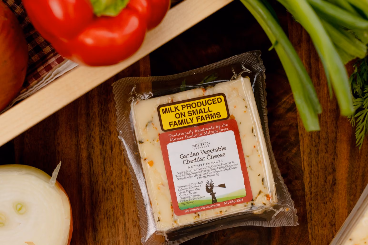 Whole and sliced yellow onions, red and green bell peppers, fresh green onions, and leafy herbs arranged on a wooden crate and cloth napkin. A larger wedge of the same cheese appears along the right edge, showing its creamy texture with visible vegetable flecks. The scene highlights farm-fresh ingredients, artisanal cheese, and a warm, handcrafted Midwest aesthetic.