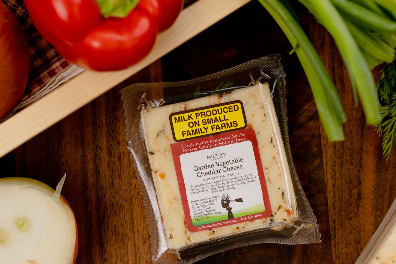 Whole and sliced yellow onions, red and green bell peppers, fresh green onions, and leafy herbs arranged on a wooden crate and cloth napkin. A larger wedge of the same cheese appears along the right edge, showing its creamy texture with visible vegetable flecks. The scene highlights farm-fresh ingredients, artisanal cheese, and a warm, handcrafted Midwest aesthetic.