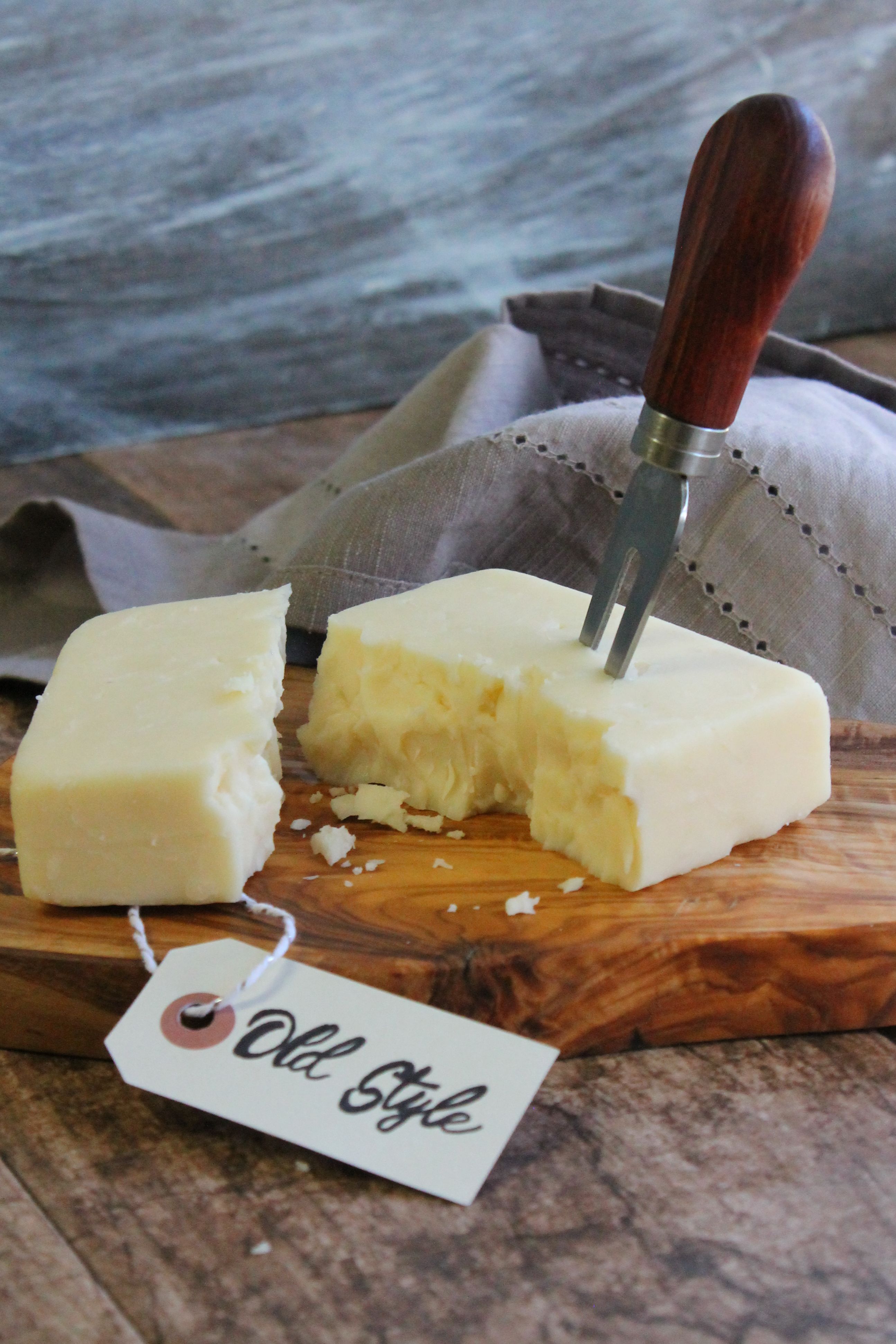 A block of cheese. One piece remains intact while the other is partially broken, with small crumbles scattered on the board, showing the cheese’s firm but delicate texture. The overall scene feels simple, traditional, and handcrafted, emphasizing the cheese’s artisanal quality and old-fashioned character.