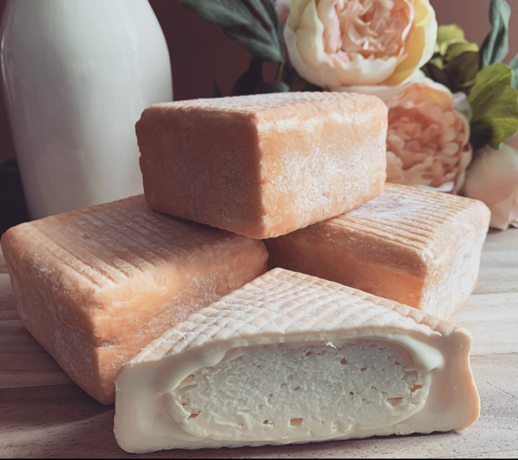 A beauty shot of our artisanal washed rind cheese cut into rectangular blocks, stacked on a wooden surface. One cheese is sliced open to reveal a creamy white interior with a soft, ripened rind. The cheeses have a pale ivory exterior with a lightly textured surface. Soft natural light, a ceramic bottle, and pale blush flowers in the background create a warm, elegant, farmhouse-style setting that highlights the craftsmanship of small-batch cheese.