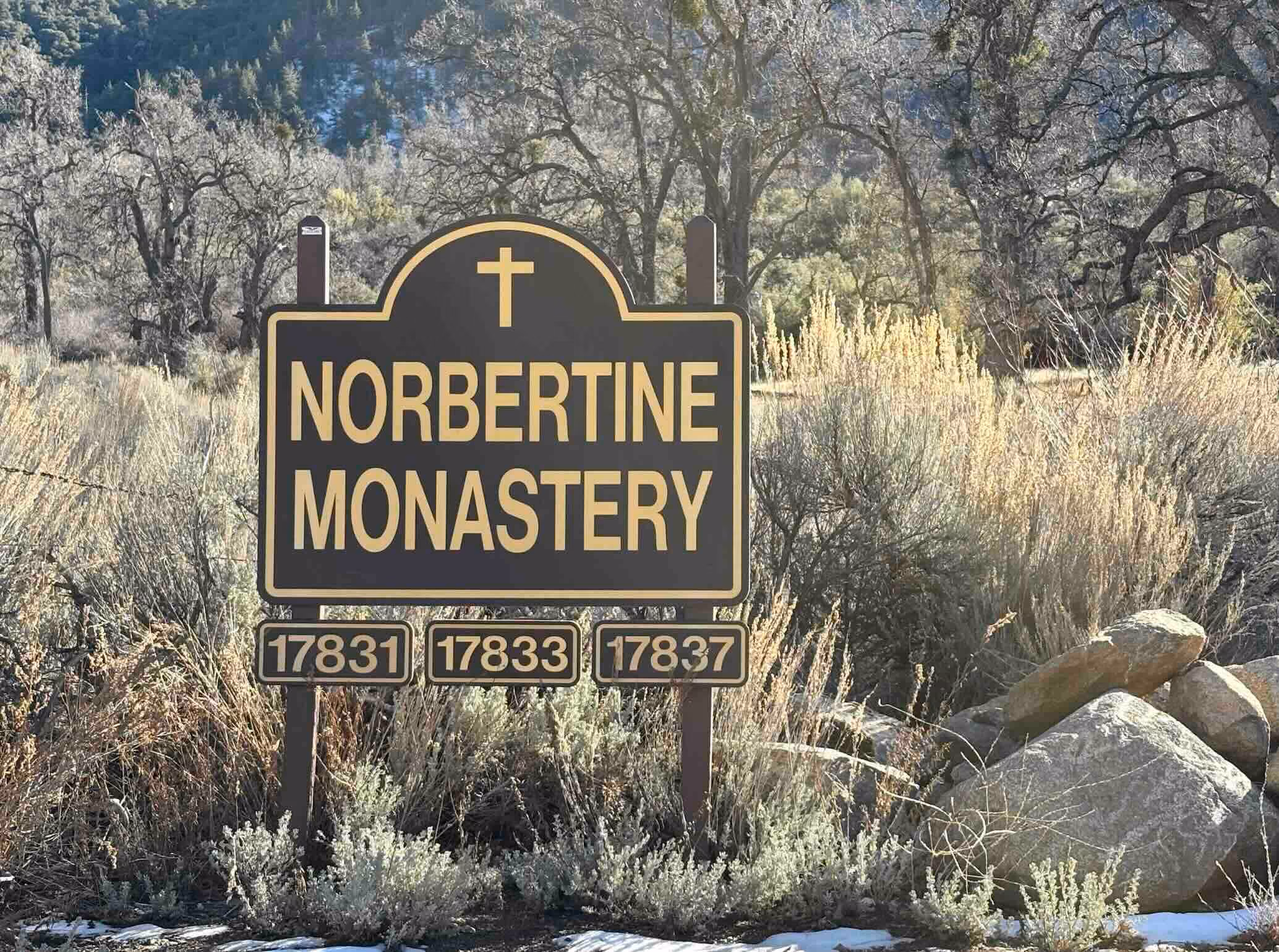 Wooden 'Norbertine Monastery' entrance sign with address numbers, surrounded by dry brush and oak trees in the Tehachapi Mountains.