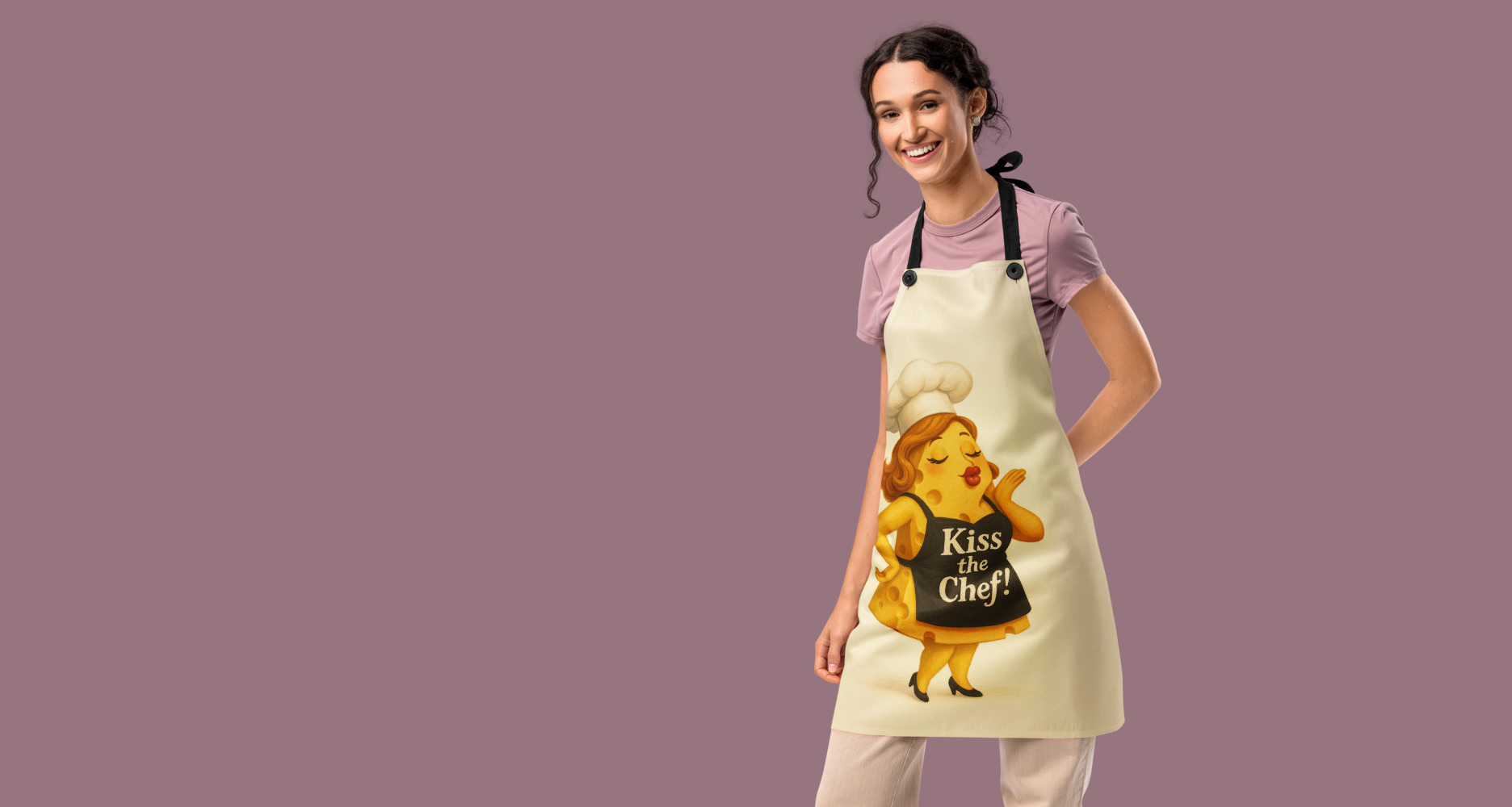 Smiling woman with braided hair against a dusty rose background, wearing a cream apron printed with a cartoon cheese character in a chef's hat blowing a kiss, lettered 'Kiss the Chef!'.