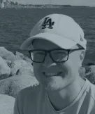 Smiling man wearing glasses and a white Los Angeles Dodgers baseball cap, with a rocky shoreline and water in the background.
