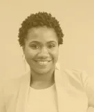 Smiling woman with short curly hair wearing a blazer and shirt against a plain background.