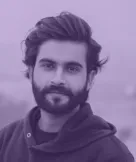 Bearded man with dark hair wearing a hoodie looking at the camera against a blurred background.