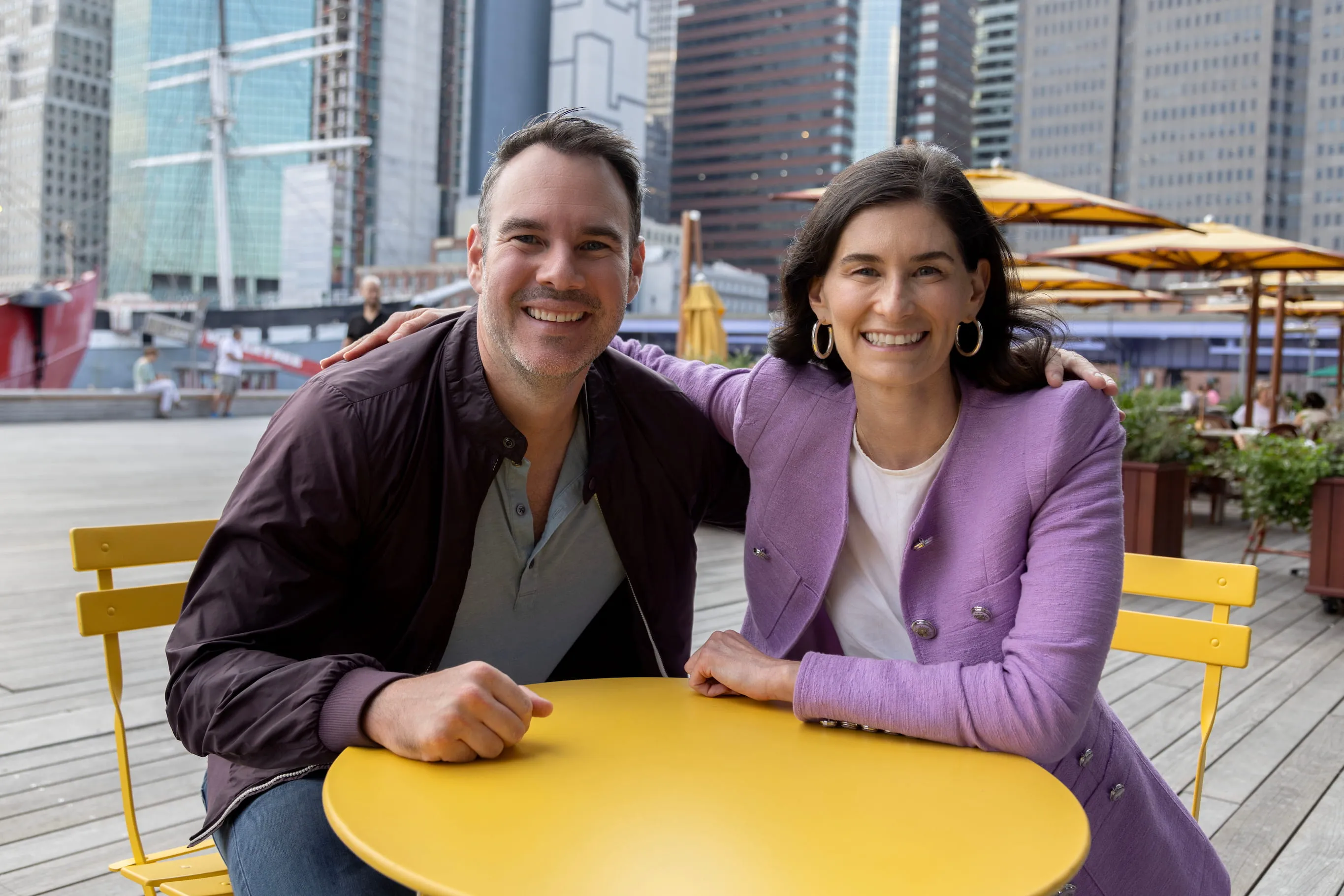 Smiling man and woman sitting closely at a yellow table in a city outdoor setting with skyscrapers in the background.