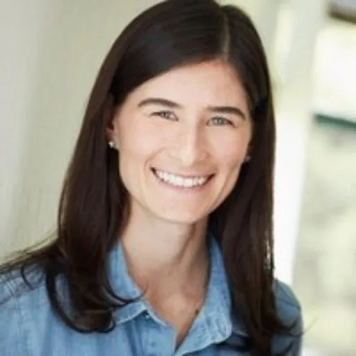 Smiling woman with long dark hair wearing a light blue shirt, in a softly lit indoor setting.