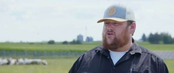 Farmer Trent Gerlach, a bearded man with a blue baseball cap and black shirt, stands with sheep and solar panels in the background.