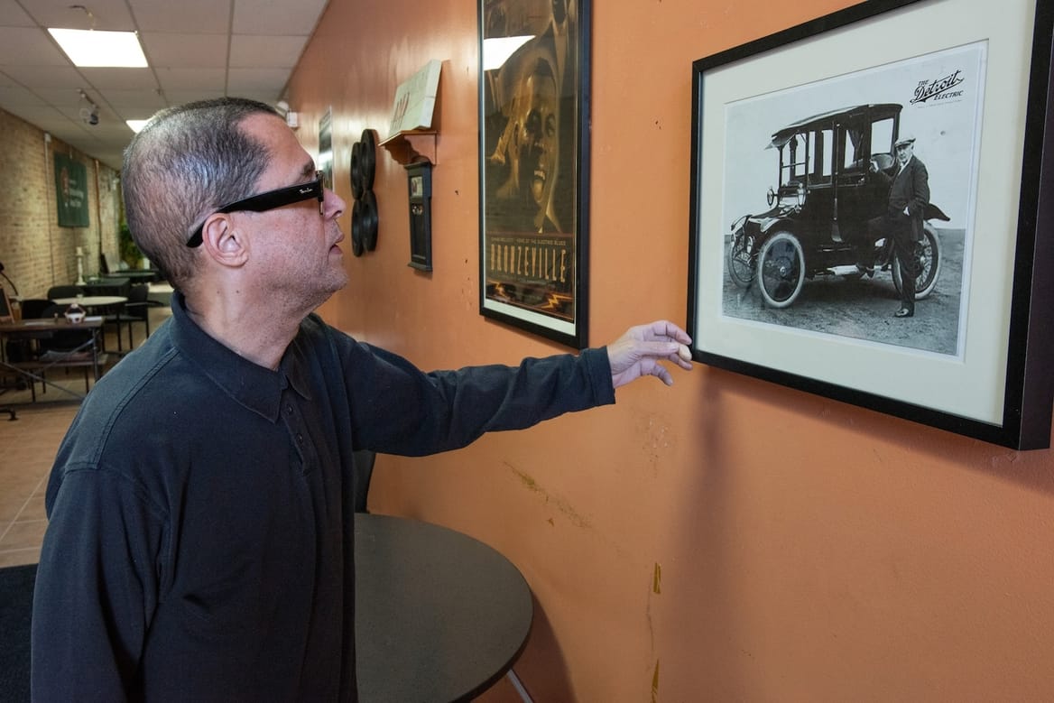 A person in sunglasses standing in a hallway reaches for a framed picture of an old-fashioned car.