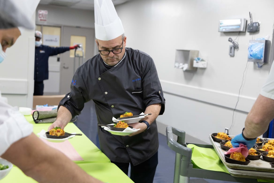 A man in glasses and a chef's uniform holds three plates of food in a commercial kitchen.