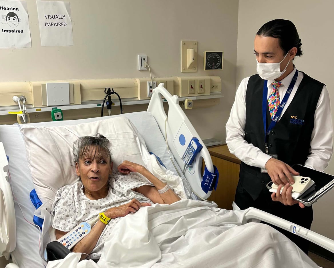 A woman lies in a hospital bed. Standing next to her is a man wearing a surgical mask and a shirt, vest, and tie.