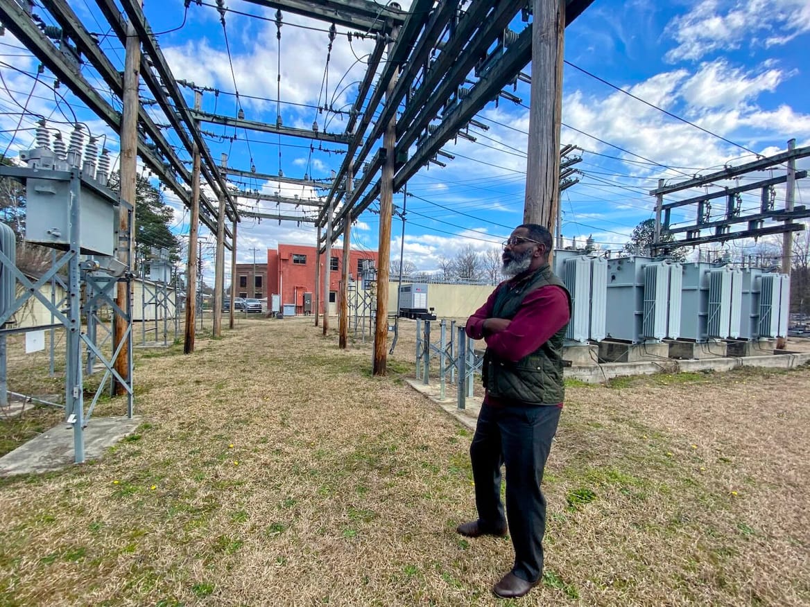 A man stands with his arms crossed and looks at electrical infrastructure, including wooden poles and wires, in a field.