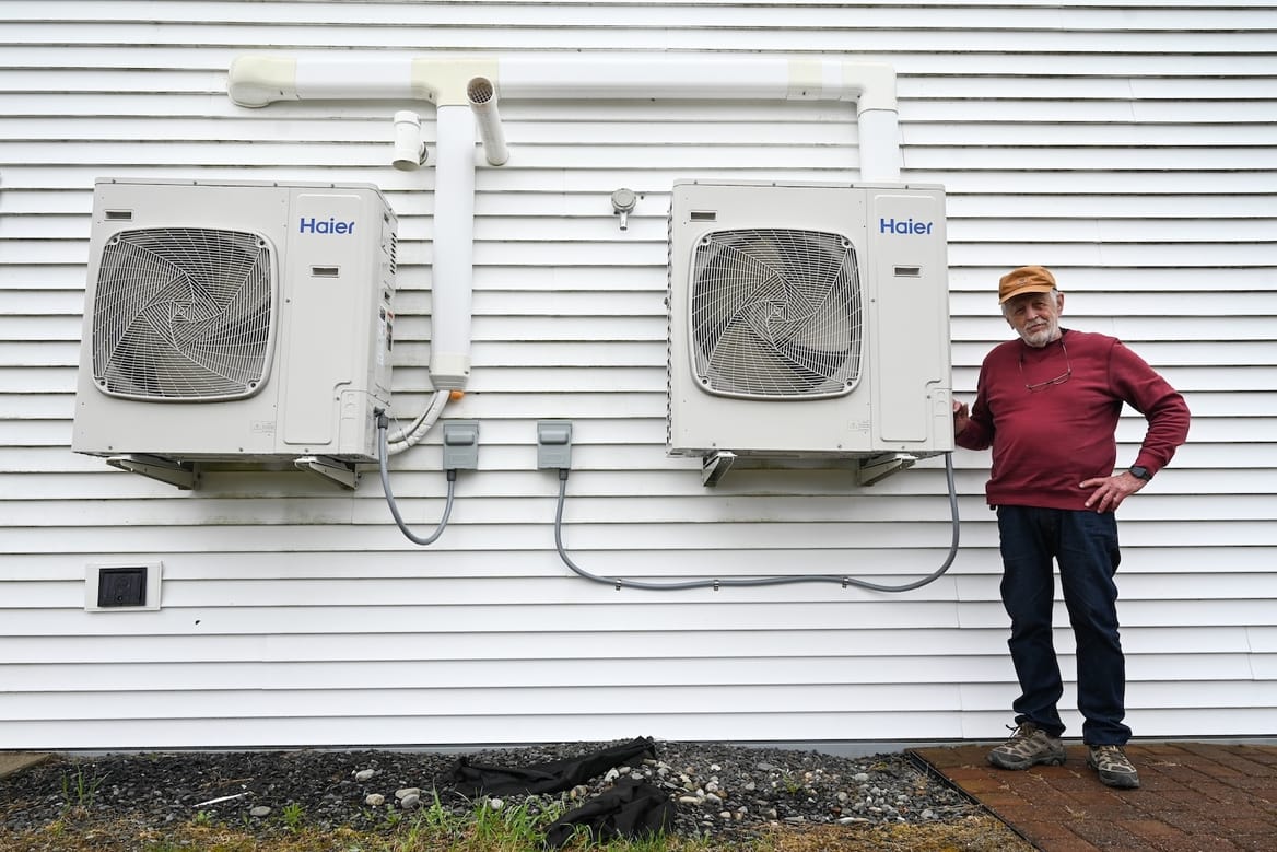 A person stands next to the exterior of a building with two heat pumps attached.
