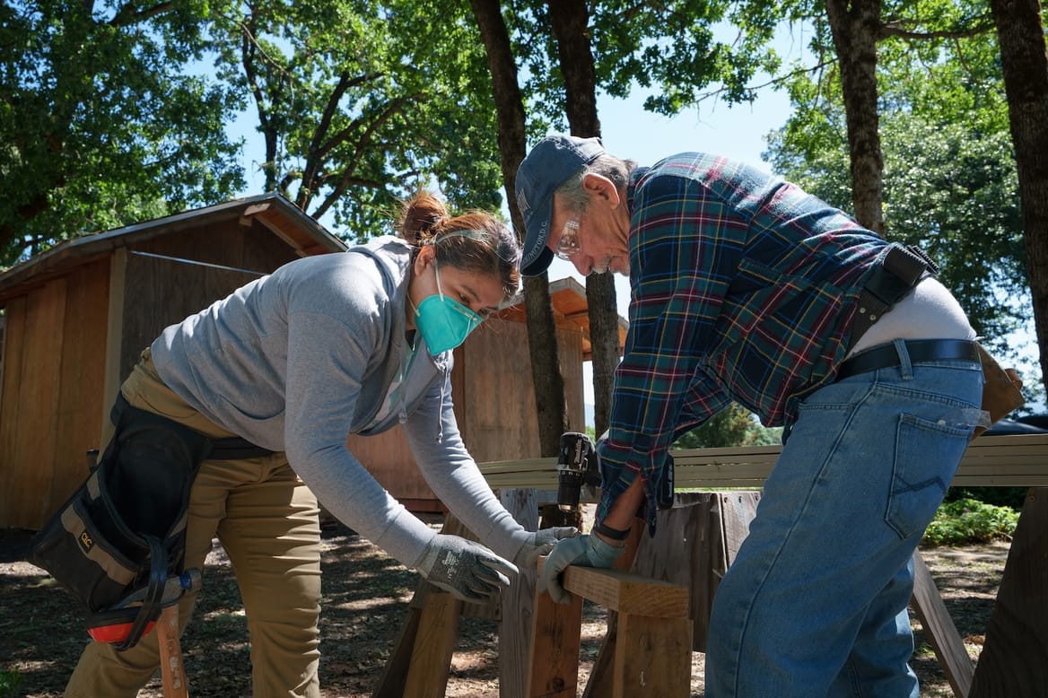 Two people wearing tool belts hold wooden beams together outside