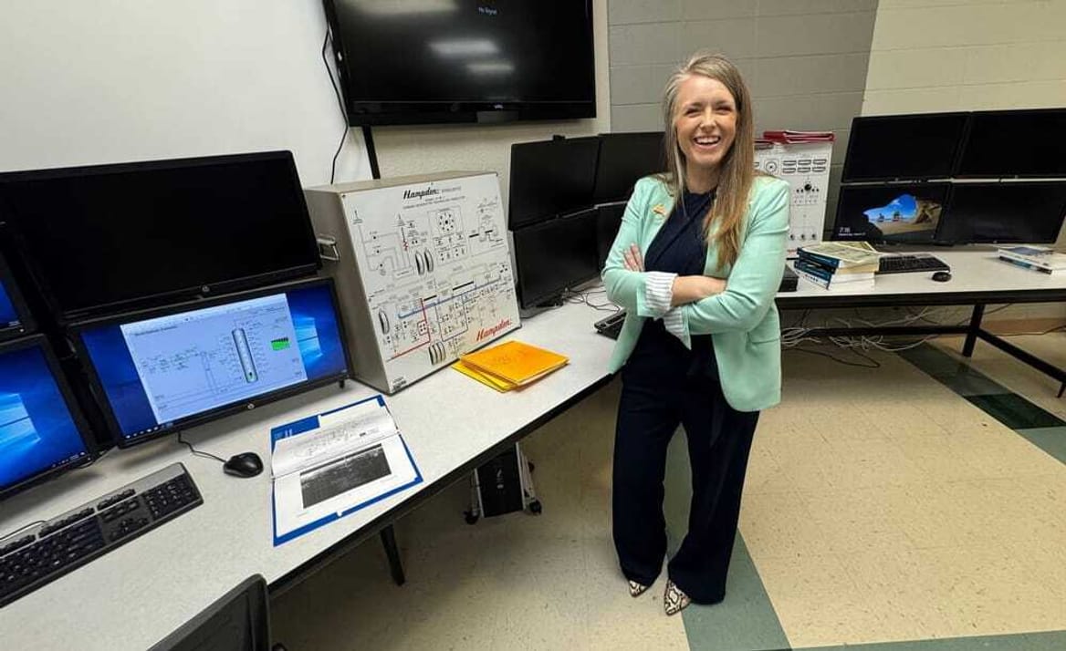 A smiling person stands next to several computers on a long desk against a wall