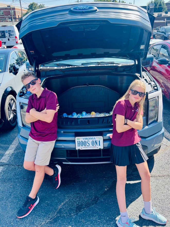 Two children stand on either side of a vehicle with an open trunk on the front end of the car