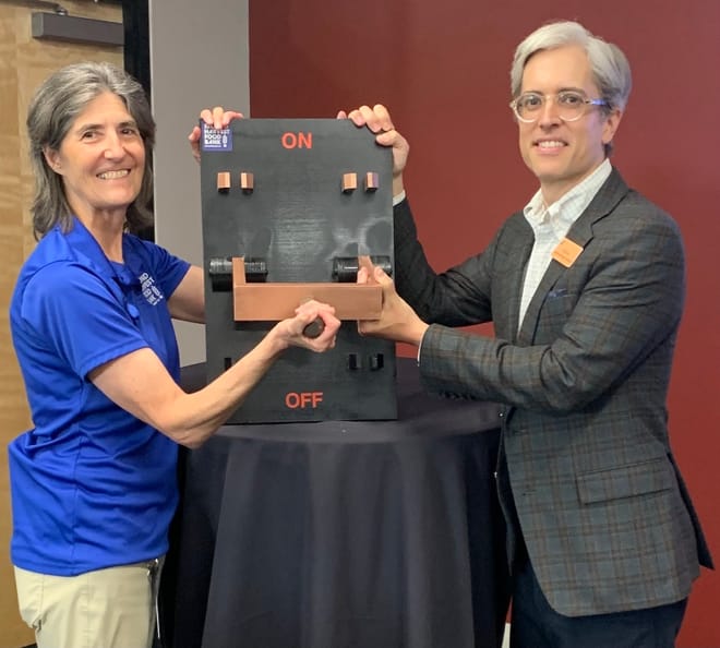 Two people pose next to an on-off switch