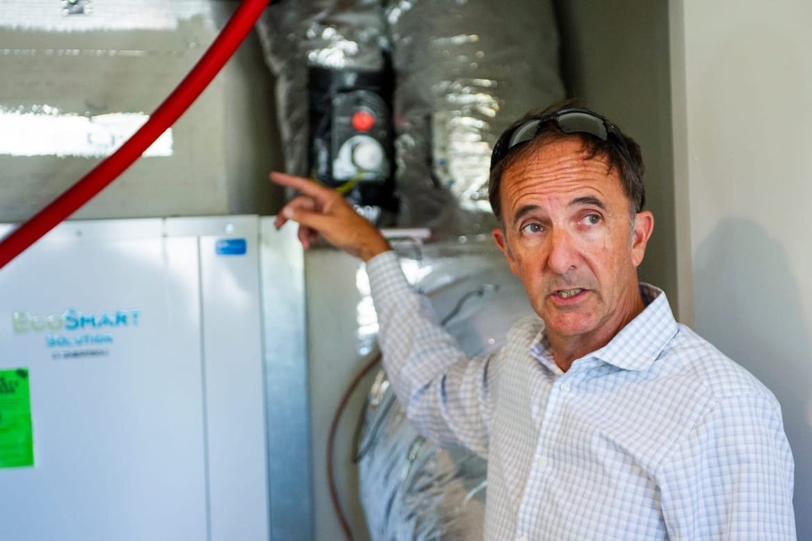 A person wearing a button-down shirt and glasses points at a white box and silver ductwork behind him