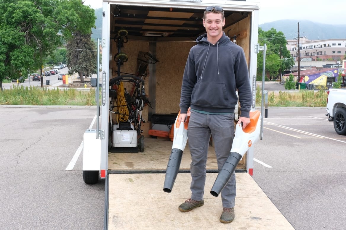 Man on equipment trailer ramp holding two electric leaf blowers.