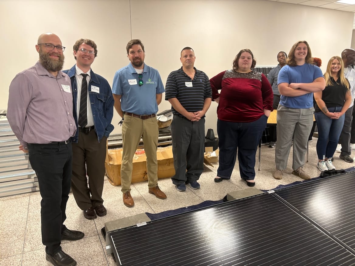 A group of people pose for an image together in a room wth a solar panel on the floor