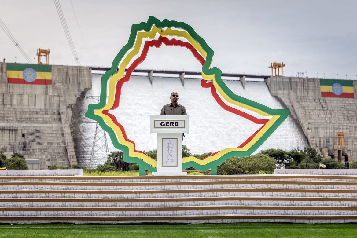 A person stands at a podium in front of a large dam
