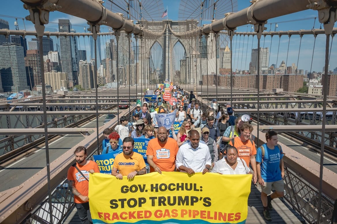 A crowd of people on a bridge near a city hold a banner reading "Gov Hochul: Stop Trump's fracked gas pipelines"