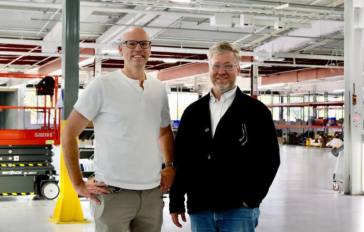 Two men stand inside a manufacturing facility with industrial equipment in the background