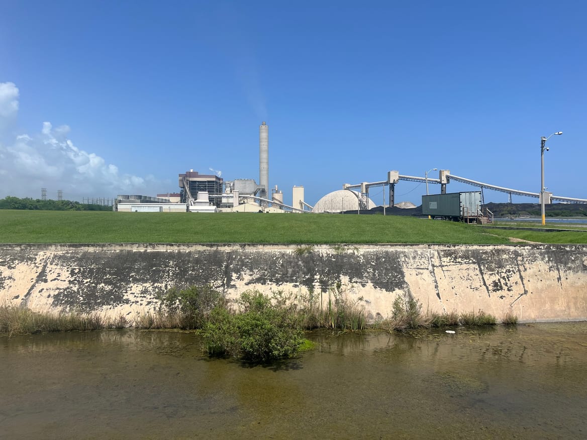The AES Puerto Rico coal plant in the distance with brown waterway and green field before it and a backdrop of blue sky