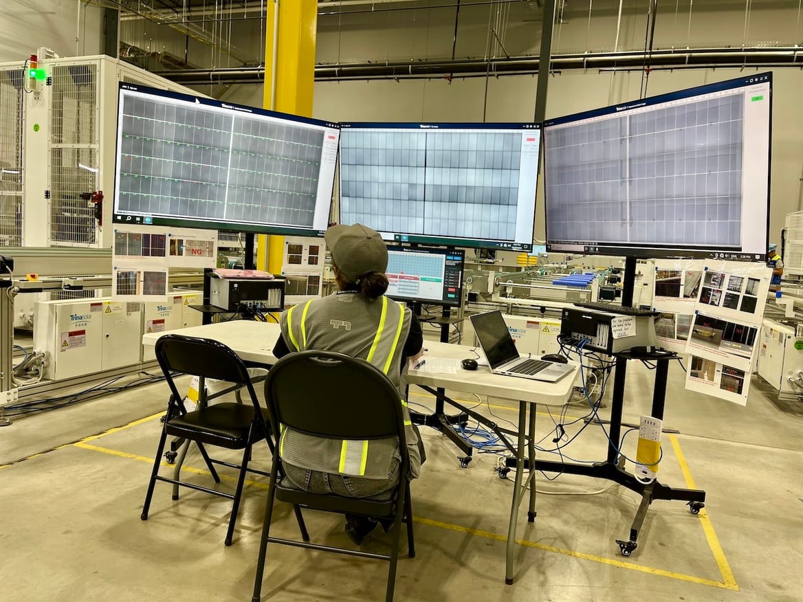 A worker, seen from the back, sits at a table with three monitors showing images of solar panels.