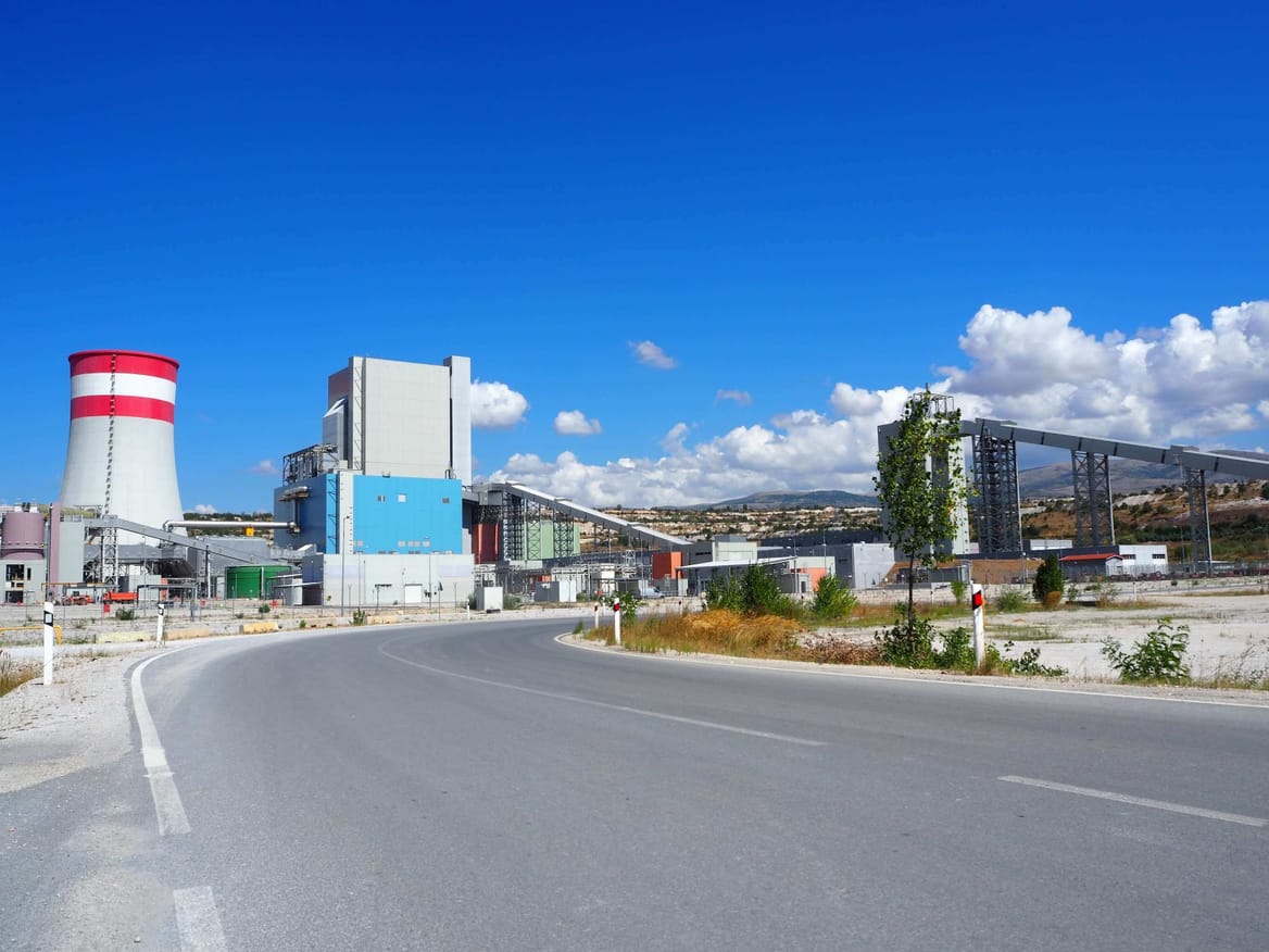 Paved road in the foreground leads to a power plant with a tower topped in red and white stripes below a blue sky