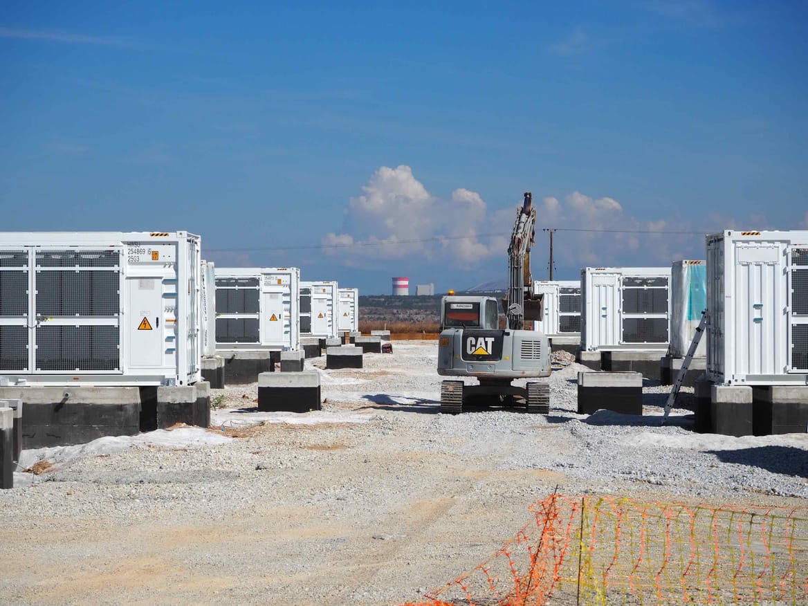 Rows of white battery containers on a dirt and stone lot with an excavator among them