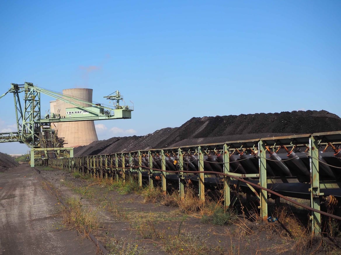 A road with piles of coal behind a green fence plus power plant equipment and towers