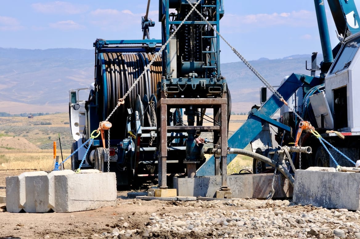 Teal blue drilling machinery bores into earth against landscape of dry grass and rolling hills.