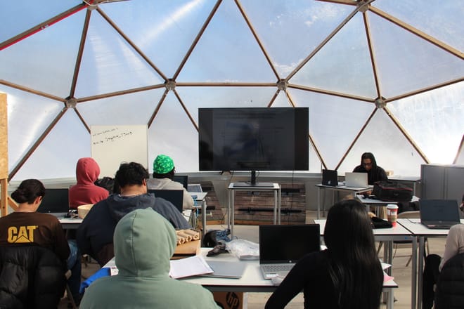 Students sit at tables facing a screen inside a geodesic dome.