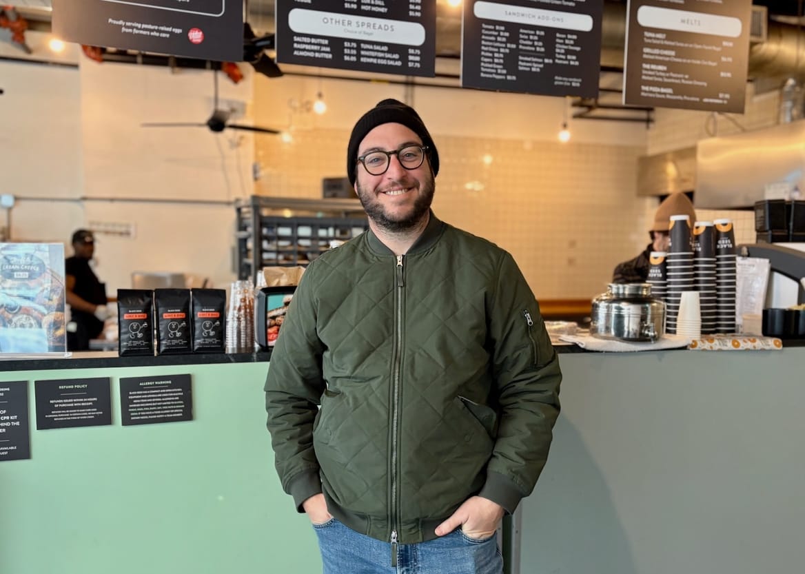 A man wearing an army green jacket and jeans, a black beanie and glasses, in front of a light green shop counter
