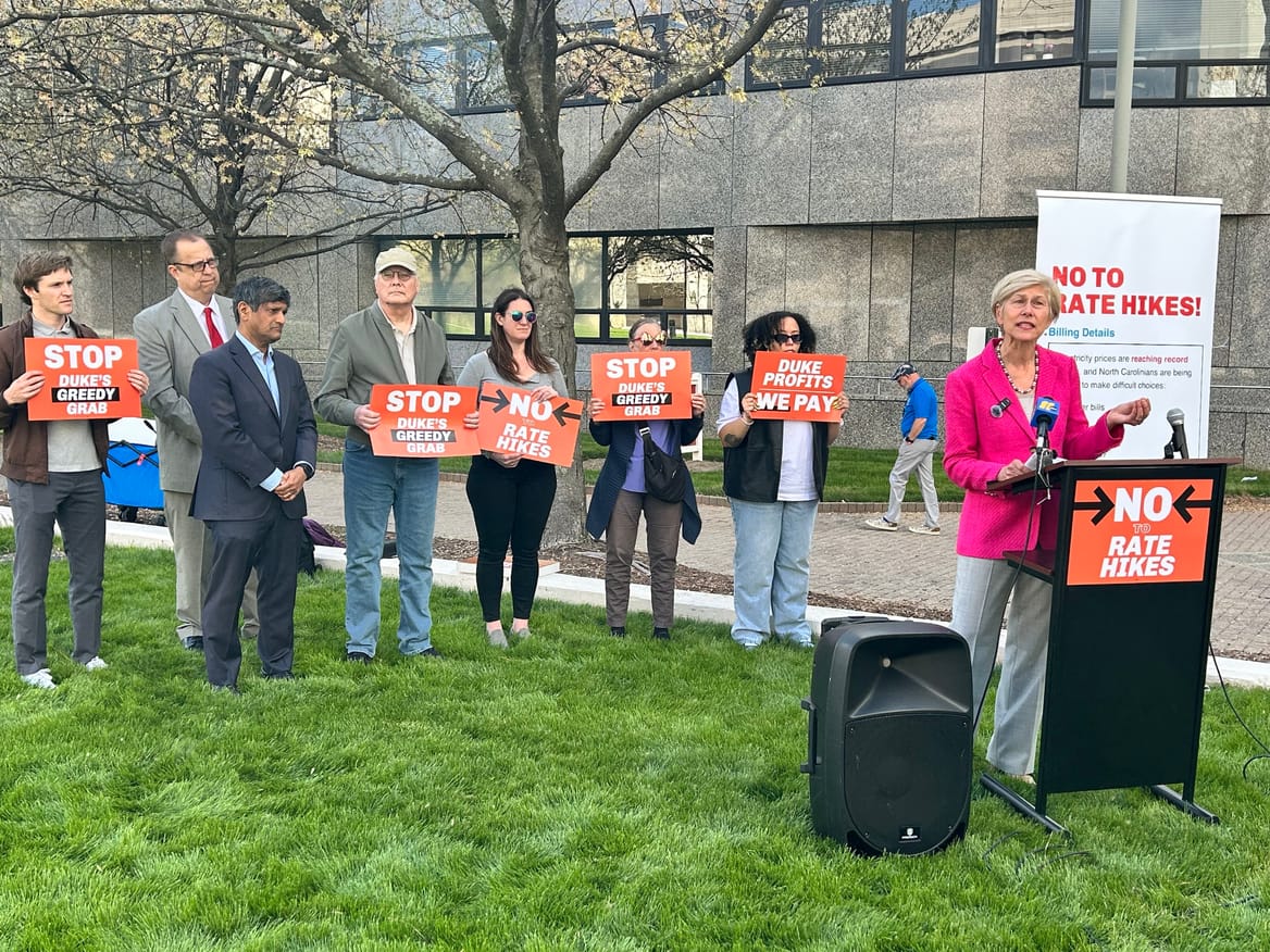 A woman in a pink blazer at a podium on a patch of grass. To her left stand 7 people, most holidng signs "No to Rate Hikes"