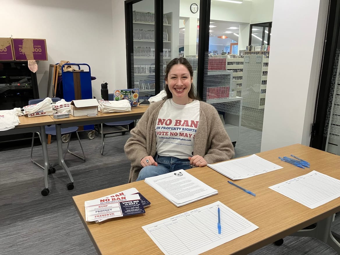A woman, wearing a "No Ban on Property Rights" T-shirt, sits at a table with flyers and forms.