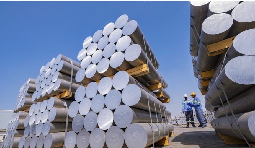 Long rolls of aluminum set on wood and stacked in groups of about 15 outside. Two workers stand between the rows