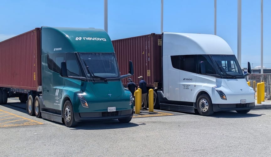 Two electric trucks, one with a green cab, the other white, with two men between them in parking lot