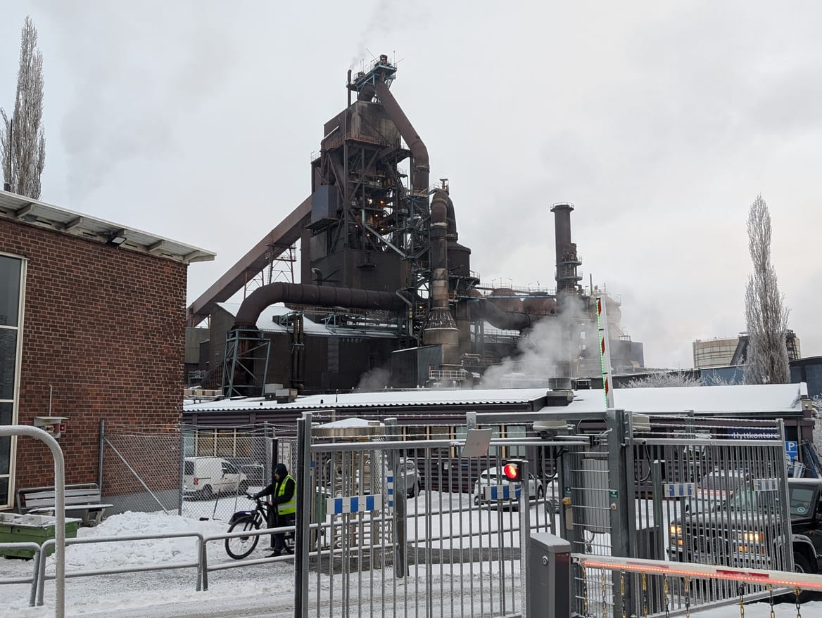 Entrance gate to steel mill site with hulking blast furnace. Snow on ground and brick building on left, plus a worker on bike