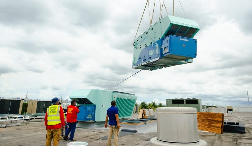 Container in shades of blue descending by crane onto a commercial building rooftop, with four people watching and guiding it